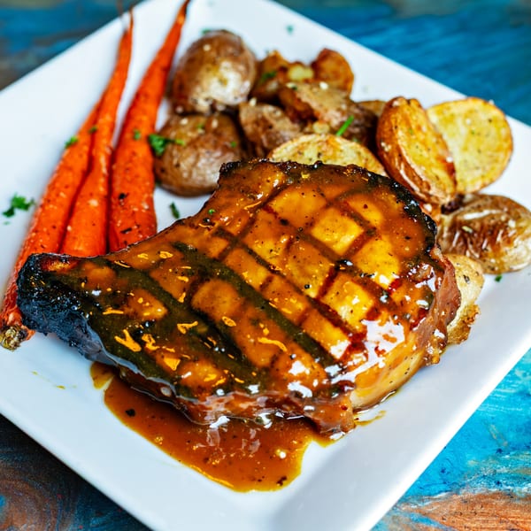 Grilled pork chop with glaze, roasted carrots, and potatoes on a white plate.