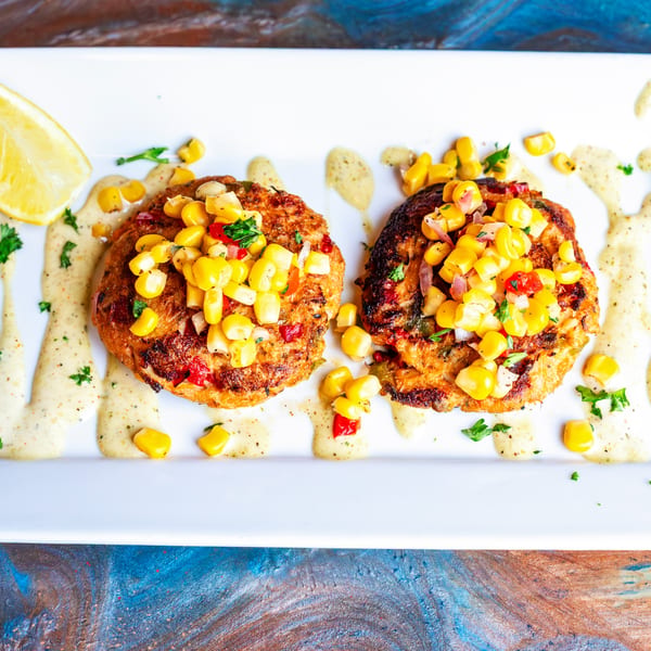 Two golden-brown crab cakes topped with corn salsa and drizzled with creamy sauce, garnished with a lemon wedge.