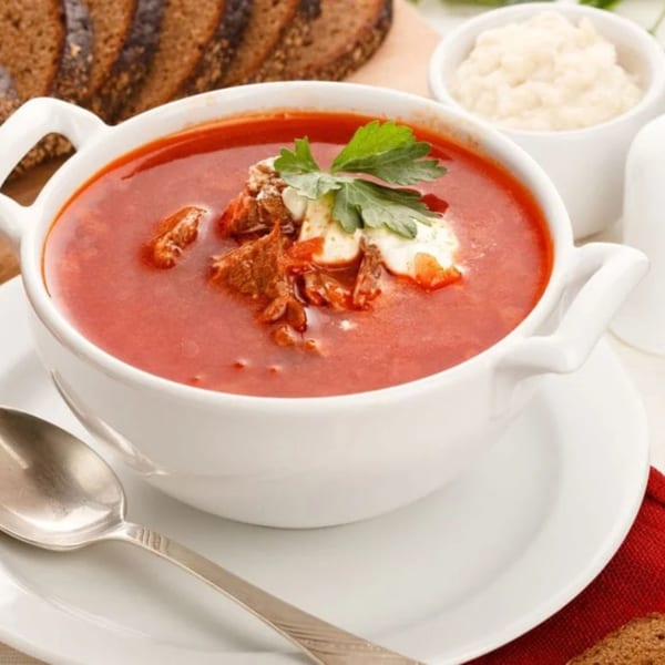 A bowl of red borscht soup with meat, sour cream, and parsley, served with dark bread.