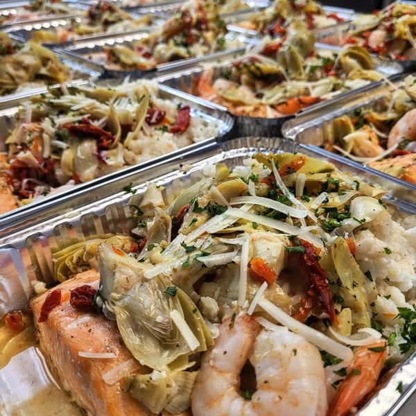Multiple foil containers filled with salmon, shrimp, artichokes, sun-dried tomatoes, and rice.